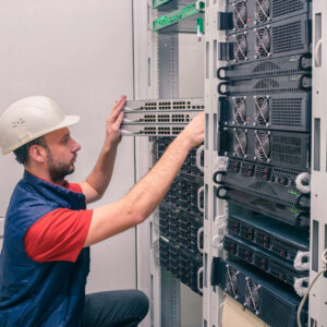 A man installs new equipment in a modern data center. An engineer in a white helmet mounts a stack of switches in a rack. The technician works in the server room.