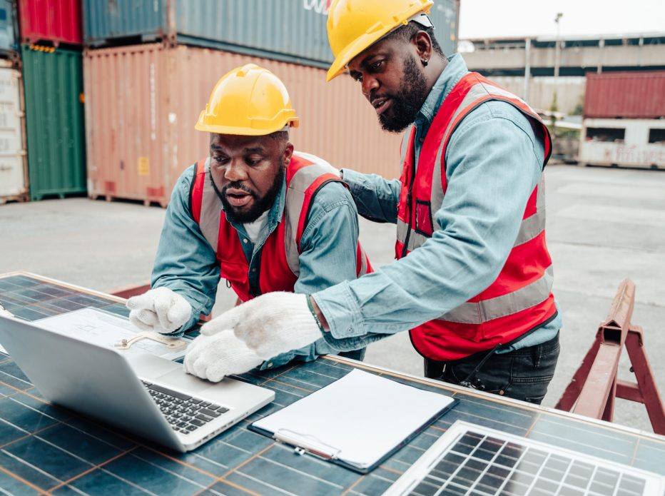Two workers in hard hats and hi-vis vests using a laptop at a shipping yard