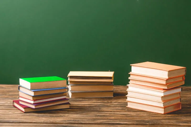 Stacks of books on a wooden desk in front of a green chalkboard
