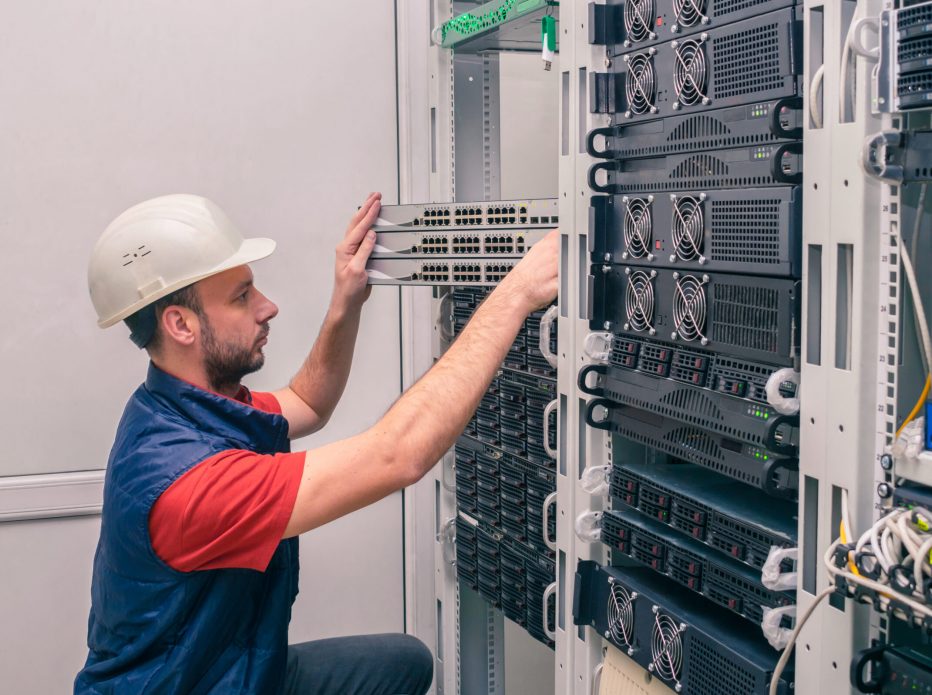 A man installs new equipment in a modern data center. An engineer in a white helmet mounts a stack of switches in a rack. The technician works in the server room.