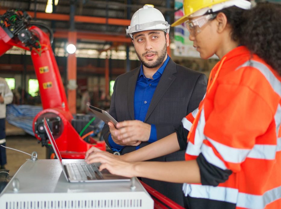 Young Africa American industrial robotic engineer talk with businessman and control robotic arm by using laptop in the factory. Manager is interested on this project. Technology and innovation concept