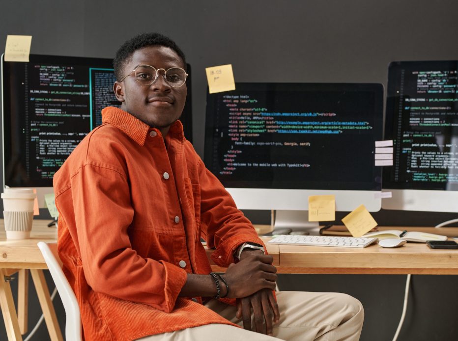 Young IT support manager looking at camera while sitting by workplace in front of computer monitors with decoded data on screens