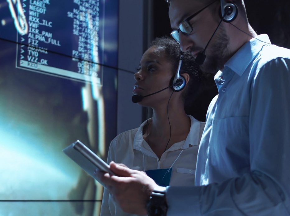 Side view of black woman and white man communicating in Moon mission control center.